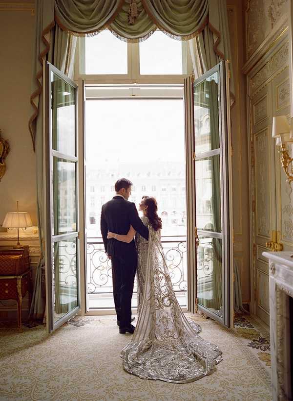 A couple portrait taken indoors in a grand Parisian hotel suite, with the bride and groom standing together facing open French doors that lead to a wrought-iron balcony overlooking a Haussmann-style cityscape. The groom wears a classic black tuxedo, while the bride wears a heavily embroidered silver and ivory long-sleeved gown with an extended cathedral train featuring intricate metallic scrollwork detail. The room features sage green draped curtains with swag valances, gold-accented moldings, a white marble fireplace, wall sconces, and a patterned cream carpet, reflecting a formal classic Parisian interior style. The image is a full-length wide shot, backlit by natural light flooding through the open doors, creating a silhouetted effect against the bright city view beyond. Potential venue feature image.