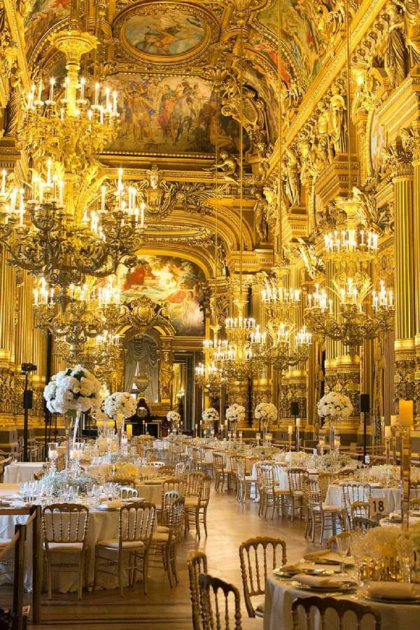 Wide shot of a wedding reception setup inside an ornate Baroque-style grand hall, identifiable as the Grand Foyer of the Palais Garnier in Paris. The room features floor-to-ceiling gilded architectural details, large painted ceiling murals, and rows of elaborate gold candelabra chandeliers casting warm amber light throughout the space. Round guest tables are dressed in white or ivory linens and set with gold Chiavari chairs, crystal glassware, and gold charger plates. Tall centerpieces of white roses and white hydrangeas on slim clear or gold pedestals are placed at regular intervals across the room, creating a consistent all-white floral scheme against the gold backdrop. No guests are present, indicating this is a pre-reception setup shot. The decor palette is white and gold with a classic formal theme. Potential venue feature image.