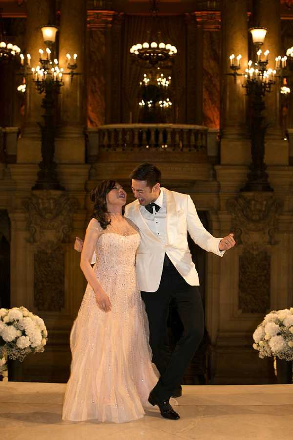 A couple shares a first dance inside a grand indoor venue featuring ornate stone balustrades, tall marble columns, and multiple candelabra-style chandeliers providing warm amber lighting. The bride wears a strapless, crystal-embellished blush gown with a fitted bodice and full skirt, while the groom is dressed in a white dinner jacket with black trousers and a black bow tie. Both are laughing and appear mid-movement on a white dance floor, with large arrangements of white hydrangeas and gypsophila flanking them on either side. The composition is a full-length portrait shot, and the architectural backdrop suggests a prestigious Parisian venue such as the Palais Garnier opera house. Potential venue feature image.