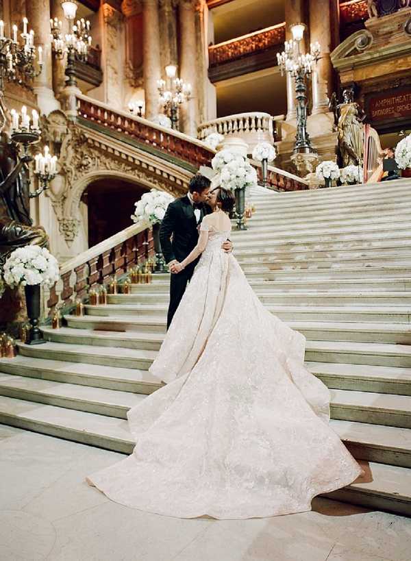 A couple shares a kiss on the grand marble staircase of the Palais Garnier (Paris Opera House), captured in a wide portrait shot from behind. The bride wears a voluminous ball gown with an extensively embroidered blush-ivory skirt and a long cathedral train that spreads dramatically across the steps, featuring off-the-shoulder detailing. The groom is dressed in a classic black tuxedo with a bow tie. The staircase is decorated with large arrangements of white flowers — likely white hydrangeas and roses — placed at intervals along the balustrades, alongside gold candle holders lining the steps. The venue's ornate gilded candelabras, arched doorways, carved stone detailing, and warm amber candlelight fill the background across multiple levels of the interior. The overall decor palette is white, gold, and ivory against the warm-toned marble architecture. Potential venue feature image.