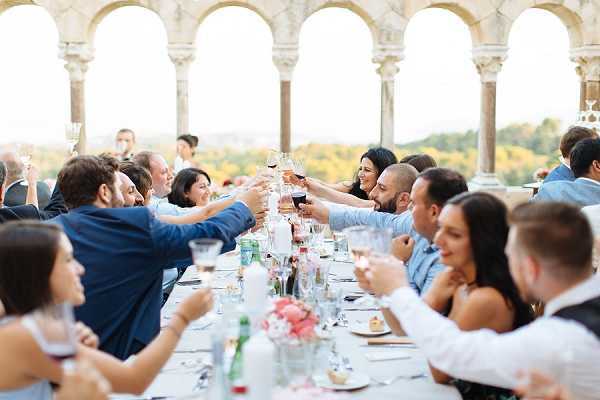 Wedding reception dinner toast captured on an outdoor terrace or loggia featuring ornate stone arched columns in the background, with approximately 20 guests seated along a long rectangular table raising their glasses in a toast. The setting appears to be an elevated historic venue with a view of greenery visible through the arches. The table is dressed with glassware, plates, and a low centerpiece of pink and white florals. Guests are dressed in smart-casual attire including navy and light blue suits and cocktail dresses. The image is a wide shot taken from the end of the table, creating depth along its length. Potential venue feature image.