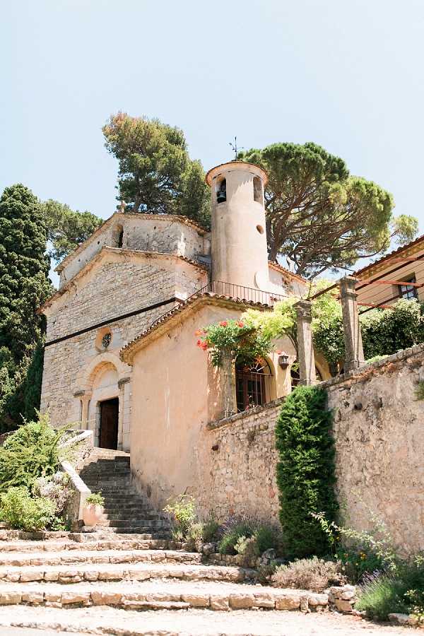 Wide exterior shot of a small Romanesque chapel with warm ochre-toned rendered walls and exposed stone detailing, featuring a distinctive round bell tower and arched doorway with stone steps leading up to the entrance. The architecture is characteristic of a historic Provençal chapel, with red flowering climbing plants trailing along the upper terrace and iron railings visible alongside an adjacent stone-walled structure. No people are present in the frame — this is a venue architectural shot taken in bright daylight. Potential venue feature image.