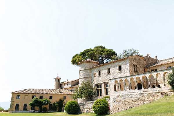 Wide exterior shot of a historic French stone chateau or manor complex, featuring a multi-story main building with arched loggia colonnades, a round tower, terracotta-tiled rooflines, and warm honey-toned limestone construction. A secondary outbuilding with matching architectural style is visible to the left. No people are present in the frame. The shot is taken from ground level on the surrounding lawn, capturing the full facade of the property in bright midday light. Potential venue feature image.