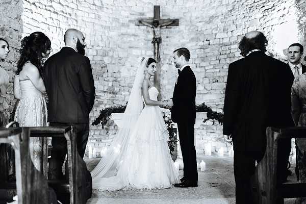 A black-and-white photograph of a wedding ceremony taking place inside a small rustic stone chapel with exposed rough-cut stone walls. The bride, wearing a strapless tiered ballgown with a long cathedral veil, and the groom, in a dark suit with a bow tie, stand facing each other and holding hands at the altar, beneath a crucifix mounted on the back wall. The altar is decorated with candles of varying heights lining both sides of the aisle and a garland of dark foliage. Several members of the bridal party and guests are visible on either side, including a bridesmaid in a light-colored dress and a man in a dark suit. The image is a wide mid-shot with strong contrast between the bright white of the bride's gown and veil and the dark stone walls, lit primarily by candlelight.