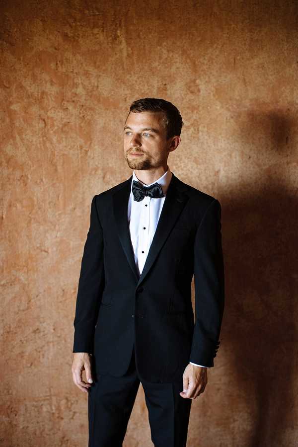 A portrait of a groom standing alone against a warm terracotta-toned textured wall, gazing slightly off-camera. He is dressed in a classic black tuxedo with satin lapels, a white dress shirt with stud buttons, and a black satin bow tie. The shot is a medium full-length portrait with natural directional lighting that casts a soft shadow across the wall behind him, highlighting the contrast between his dark tuxedo and the warm background tones.