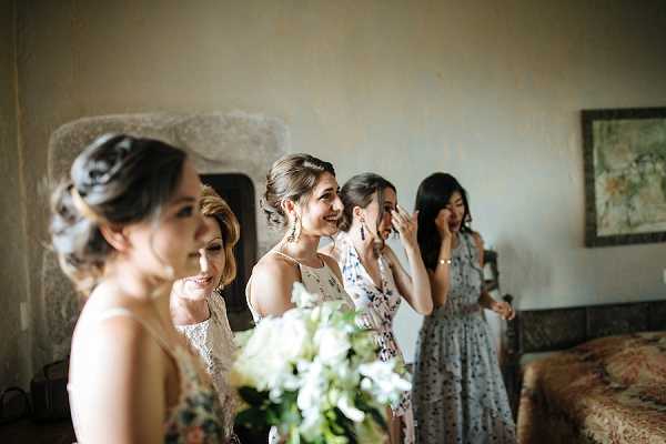 An indoor getting-ready moment captures a group of four women reacting emotionally — two are visibly wiping tears from their eyes — likely upon seeing the bride for the first time. The setting appears to be a room with aged plaster walls, a framed artwork, and a patterned upholstered chair, suggesting a chateau or historic venue interior. The women wear varied outfits including a white lace top, a floral printed dress, and a muted blue-grey floral midi dress, with their hair styled in upswept arrangements. In the foreground, a partial view of a white and green bouquet featuring what appear to be white ranunculus or garden roses with lush greenery is visible. The composition is a candid medium shot with natural window light, capturing a genuine emotional reaction during the pre-ceremony preparations.
