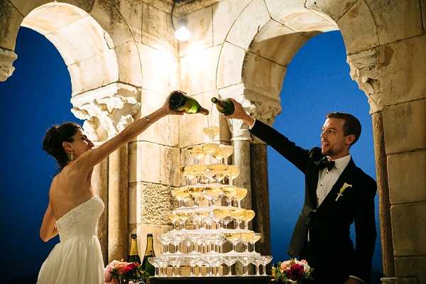 A bride and groom are pouring champagne simultaneously into the top of a champagne tower, with the liquid cascading down through multiple tiers of coupe glasses. The setting is an outdoor stone arcade or cloister with carved archways and columns, lit by a warm overhead spotlight against a deep blue evening sky. The bride wears a strapless white gown and the groom is dressed in a dark navy tuxedo with a black bow tie and a white boutonniere. The table holding the champagne tower is decorated with pink and red floral arrangements, and an additional champagne bottle is visible on the table. The image is a wide portrait shot capturing both subjects and the full height of the tower.