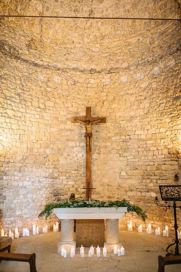 Detail shot of a chapel altar decorated for a wedding ceremony. The altar is a simple stone table adorned with a trailing greenery garland, likely eucalyptus or ivy, running along its top edge. A wooden crucifix with a gold-toned corpus stands prominently behind the altar against a curved apse wall of exposed pale limestone. Dozens of white pillar and votive candles are arranged in clusters across the stone floor surrounding the altar, providing warm ambient lighting. A wrought-iron music or lectern stand is visible to the right. The overall decor palette is minimal — white candles and green foliage against the warm stone — suggesting a classic, understated religious ceremony style. Potential venue feature image.