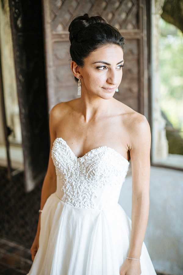 A bridal portrait of a single bride shot from approximately the waist up, set against a backdrop of aged wooden architectural columns or doors, likely at a chateau or historic venue. The bride wears a strapless white ball gown with a heavily beaded and embroidered sweetheart bodice, paired with long crystal drop earrings and a delicate bracelet. Her dark hair is styled in a high, sleek updo, and she is gazing off to one side. The composition is a close-up portrait with soft natural light, and the overall styling is classic and polished.