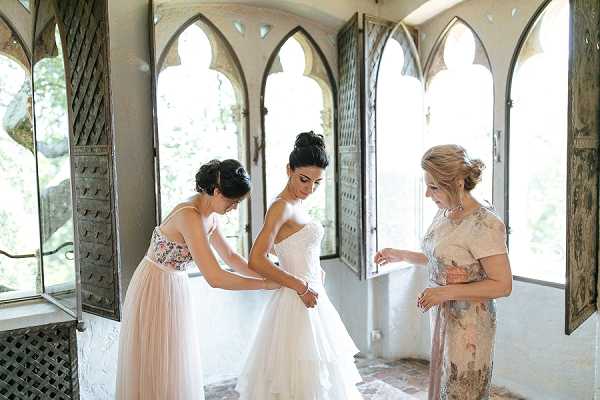 A bride is being helped into her strapless ivory ballgown with a full tulle skirt inside a room with distinctive Gothic-arched windows featuring ornate carved wooden shutters in a weathered grey finish. Two women assist her — one on the left wearing a blush tulle skirt with a floral-print top, and one on the right in a floral jacquard dress with short sleeves in muted blush and gold tones. The interior space has white plaster walls and the arched windows allow soft natural light to fill the room. This is a getting-ready portrait shot taken at medium range, capturing all three figures from approximately knee height upward.