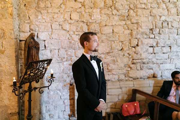 The groom stands at the altar of a stone chapel, facing forward with his hands clasped in front of him as he waits during the ceremony. He is wearing a classic black tuxedo with a white dress shirt, black bow tie, and a white boutonniere on his lapel. The interior features exposed limestone block walls, a wrought iron candelabra with lit candles, and a small religious statue mounted on the wall, giving the space a historic chapel aesthetic. A male guest is partially visible in the wooden pews to the right in the mid-ground; portrait shot with a shallow depth of field.
