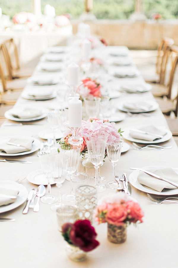 A close-up detail shot of a long reception dining table set for a wedding meal, photographed from one end with a shallow depth of field. The table is dressed with a white linen and set with white plates, silver flatware, and folded grey linen napkins, accompanied by crystal glassware in varied heights. Centerpieces along the table runner include low arrangements of blush pink hydrangeas and coral garden roses, with smaller clusters of deep burgundy roses and additional coral blooms in metallic vessels near the foreground, alongside white pillar candles and small latticed votive holders. The overall decor palette is white, blush, coral, and burgundy with a classic, romantic styling; natural wood crossback chairs are visible on either side, and the setting appears to be under an open-sided covered terrace with greenery visible in the background.