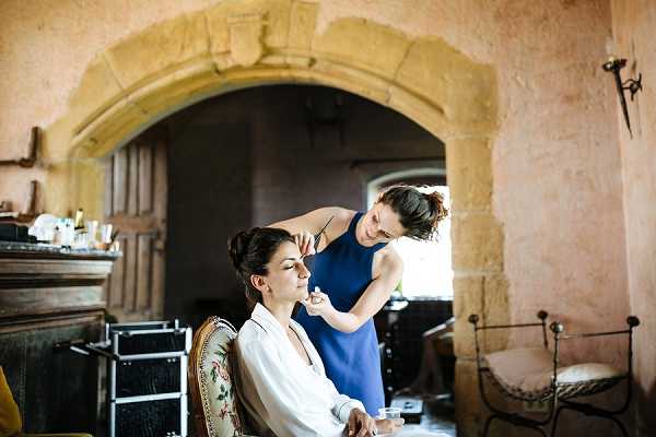 A bride is having her makeup applied during the getting-ready portion of the wedding day, seated in an ornate upholstered chair with a floral pattern. She is wearing a white silk or satin robe and has her dark hair styled up, while a makeup artist in a navy blue sleeveless top works on her face with a brush. The setting is a historic interior space with warm terracotta-toned plastered walls and a large stone archway with carved detailing visible in the background, consistent with a French chateau or manor house. Professional makeup kit cases and product bottles are visible to the left, and a wrought iron bed frame is partially visible to the right. The shot is a medium portrait taken at eye level.