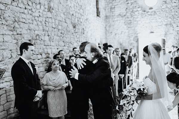 A black-and-white photograph capturing an emotional moment during a wedding ceremony inside a stone chapel, where the groom embraces a religious officiant or family member near the altar. The bride stands to the right, smiling and holding a large cascading bouquet of flowers, wearing a white gown and a long veil. A groomsman in a dark suit stands to the left, and a full congregation of seated and standing guests is visible in the background. The image is a mid-range candid shot with soft contrast, emphasizing the emotional exchange between the groom and the other man.