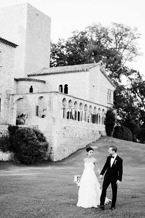 This black-and-white couple portrait shows a bride and groom walking hand-in-hand across a lawn in front of a large historic stone building with Romanesque-style arched colonnades, a tower, and tiered terraces. The bride wears a strapless ballgown with a layered skirt and carries a bouquet, while the groom is dressed in a dark tuxedo with a bow tie and boutonniere. The image is shot in a wide-angle, full-length style with high contrast tones, placing the couple in the lower foreground against the dramatic scale of the venue architecture. Guests and what appears to be a reception setup are faintly visible on one of the terraces in the background. Potential venue feature image.