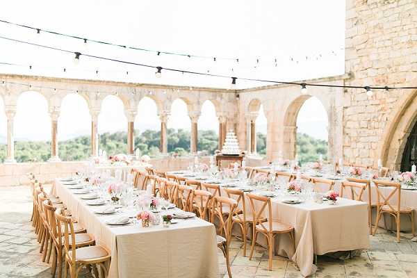 Outdoor wedding reception set up on a stone terrace of a historic château or abbey, featuring a colonnade of rounded arches with views of the surrounding landscape beyond. Three long rectangular banquet tables are dressed in blush-pink linen tablecloths and set with glassware, charger plates, and low centerpieces of pink and coral florals in small vases. Natural wood cross-back chairs line both sides of each table. A multi-tiered white wedding cake is displayed on a wooden stand at the far end between the tables. String bistro lights are strung overhead across the open-air terrace, adding ambient lighting. The overall decor palette is blush pink and warm natural wood with a classic, romantic style. Wide establishing shot capturing the full table layout and architectural setting. Potential venue feature image.