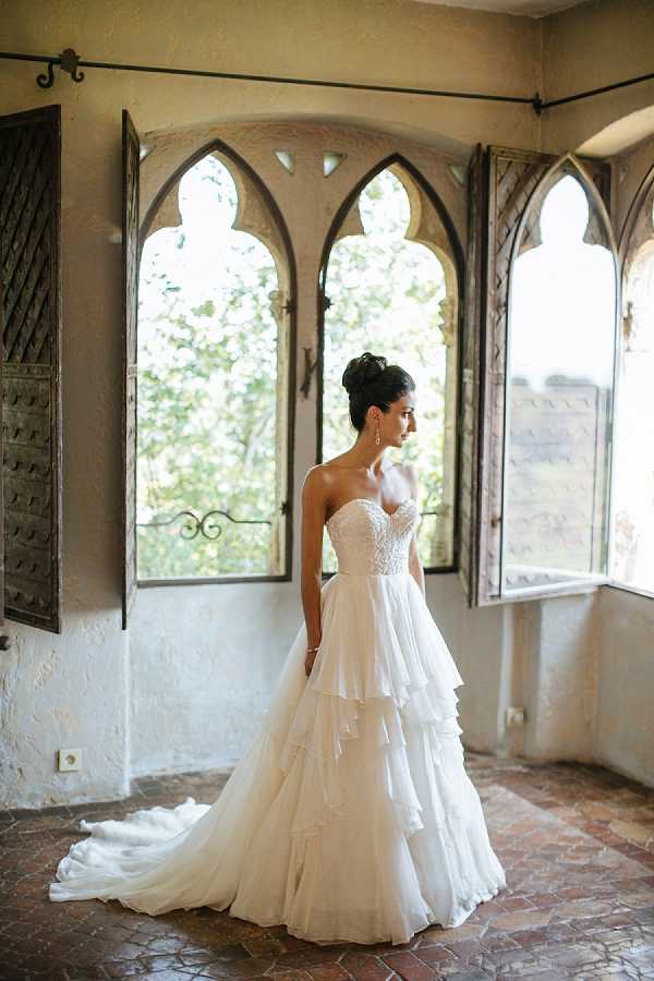 A bridal portrait of a single bride standing indoors in front of a row of Gothic-arched windows with aged wooden shutters, likely inside a chateau or historic venue. The bride wears a strapless ivory ball gown with a lace-embellished sweetheart bodice and a multi-tiered layered tulle skirt with a cathedral train, paired with drop earrings and her dark hair styled in an updo. The room features terracotta tile flooring and textured plaster walls, giving it a classic, historic character. The shot is a full-length portrait with natural backlight flooding through the arched windows.