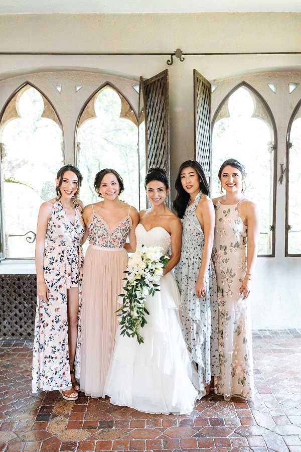 A bridal party portrait featuring the bride and three bridesmaids posing indoors in front of gothic arch windows with wooden shutters. The bride wears a strapless white ball gown with a lace bodice and carries a cascading bouquet of white blooms and greenery. The bridesmaids wear mismatched floor-length dresses in a coordinated mix-and-match style: one in a blush floral print, one in a blush two-piece with a beaded top and tulle skirt, and one in a pale grey-blue floral print, and one in a champagne floral embroidered gown. The setting appears to be a Mediterranean or Spanish Colonial-style interior with terracotta tile flooring and ornate arched window frames, giving the image a classic romantic feel. This is a medium-distance portrait shot with natural light filtering through the windows behind the group.