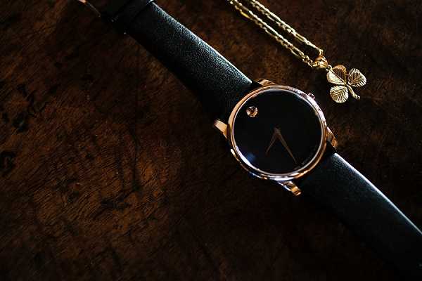 A close-up detail shot of wedding accessories laid flat on a dark wood surface. A minimalist watch with a black dial, gold-tone case, and black leather strap — consistent with a Movado Museum-style design — is placed alongside a delicate gold chain necklace with a four-leaf clover pendant charm. The image is shot with dramatic, low-key lighting that emphasizes the contrast between the gold hardware and the dark background, creating deep shadows across the wooden surface. This appears to be a groom or groomsman accessory flat-lay detail shot.