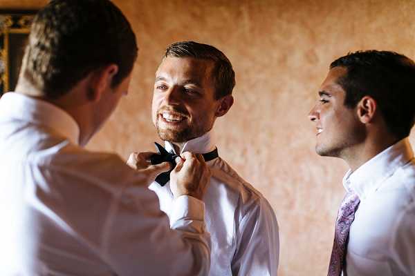 A getting-ready moment indoors showing three men, with one groomsman adjusting a black bow tie on the groom while a second groomsman looks on smiling. The groom is wearing a white dress shirt and black bow tie, one groomsman wears a white shirt, and the other wears a white shirt with a light lavender paisley tie. The setting appears to be an interior room with warm, textured ochre-toned walls. The image is a close-up portrait shot with soft, warm lighting.