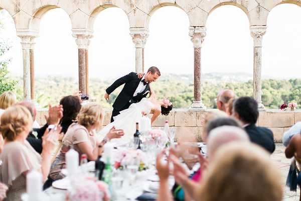The couple shares a dramatic dip-kiss moment during what appears to be their first dance or a celebratory moment at the reception, with the groom in a black tuxedo lifting and dipping the bride in a white gown while guests seated at long banquet tables on either side applaud and react with enthusiasm. The setting is an outdoor terrace or loggia of a historic stone venue featuring a row of ornate Romanesque arched columns and carved capitals, with a tree-lined landscape visible in the background. Guest tables are dressed with white linens and feature low pink floral centerpieces, glassware, and wine bottles. The shot is a wide, candid reception photograph that places the couple centrally between the tables, framed by the architectural colonnade behind them.