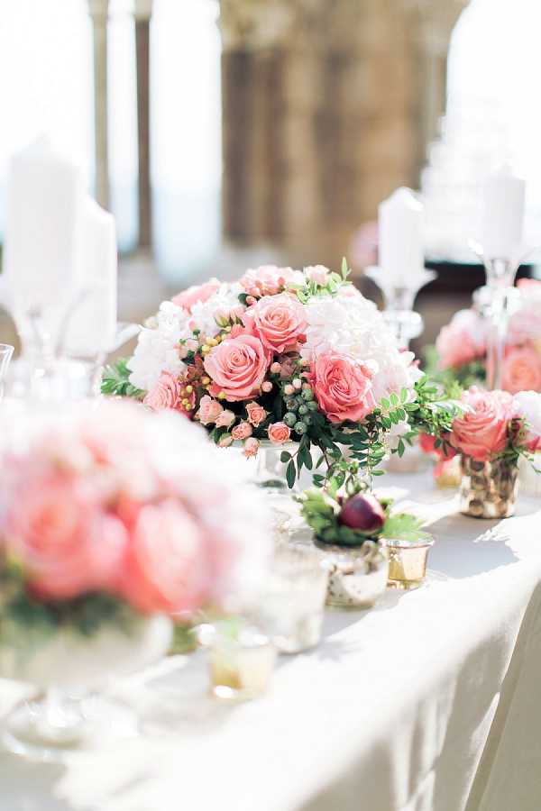 Close-up detail shot of a reception tablescape featuring low floral centerpieces arranged along a white linen-covered table in what appears to be an ornate indoor venue. The centerpieces are composed of pink roses in varying shades from blush to coral, white hydrangeas, small spray roses, green berries, and eucalyptus foliage, displayed in small silver and gold mercury glass vessels. Gold votive candle holders and crystal glassware are scattered along the table, alongside tall white pillar candles in the background. The overall decor palette is blush pink, white, and gold with a classic, romantic styling. The image is shot with a shallow depth of field, keeping the foreground and background softly blurred while the central arrangement is in focus.