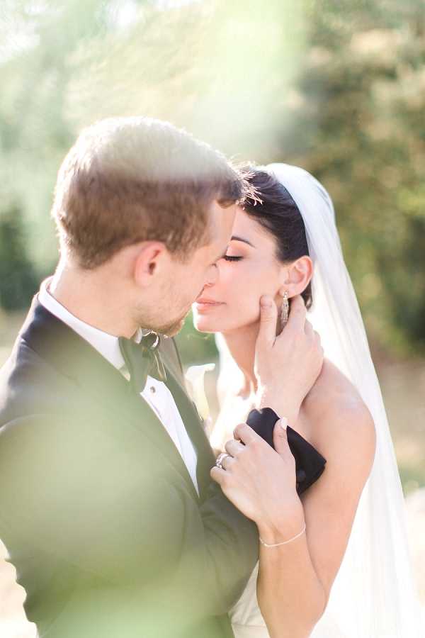 A close-up portrait of a bride and groom sharing an intimate moment outdoors, with the groom gently cradling the bride's face as they lean in close together. The groom wears a dark navy or charcoal suit with a white dress shirt and an olive or sage green bow tie, while the bride wears a strapless white gown and a plain cathedral-length veil with her dark hair styled up, accessorized with drop earrings and a delicate bracelet. The bride's engagement and wedding rings are visible on her hand resting on the groom's lapel. The image is shot with a very shallow depth of field, creating a soft, backlit, high-key effect with lens flare softening the edges of the frame.