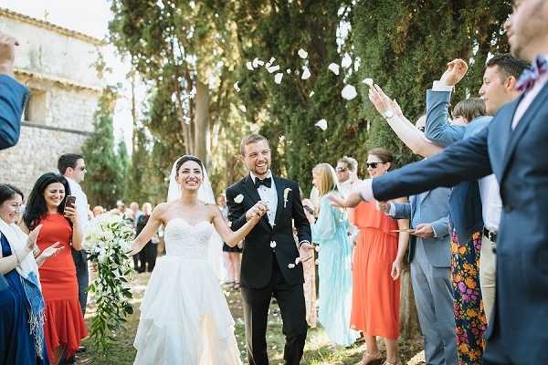A bride and groom walk hand-in-hand through a guest-lined aisle as white flower petals are tossed in the air around them during an outdoor ceremony exit. The setting appears to be a stone chapel or church exterior in the South of France, with tall cypress trees visible in the background. The bride wears a strapless, sweetheart-neckline ballgown with a layered tulle skirt and a cathedral veil, carrying a loose greenery-forward bouquet with white blooms and trailing eucalyptus; the groom is dressed in a black tuxedo with a white boutonnière and bow tie. Approximately 15–20 guests line both sides, dressed in colorful summer attire including a red dress and a floral-print dress, tossing petals while smiling and photographing the moment — captured as a medium-wide portrait shot at golden hour.
