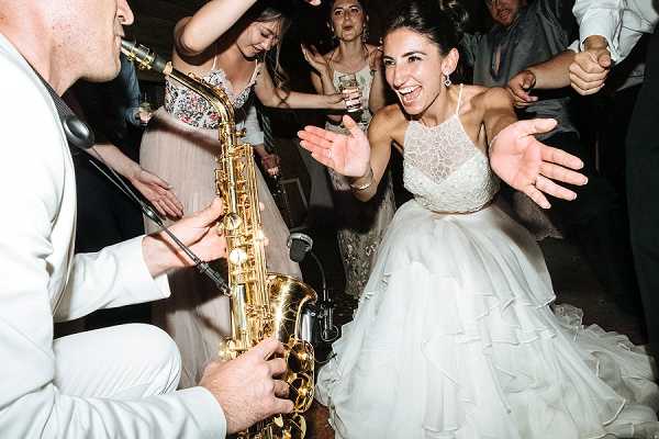 A lively reception dance floor scene captured in a close-up, dynamic shot. The bride, wearing a white layered ballgown with a halter-neck lace bodice, crouches toward a saxophonist dressed in a white suit who is playing a gold alto saxophone directly in front of her. Her expression is one of pure excitement, with hands spread wide. Several guests are visible dancing in the background, including a woman in a blush floral dress and another in a silver-toned gown, all appearing animated and energetic. The setting appears to be an indoor reception venue with dim, warm lighting typical of an evening dance floor.
