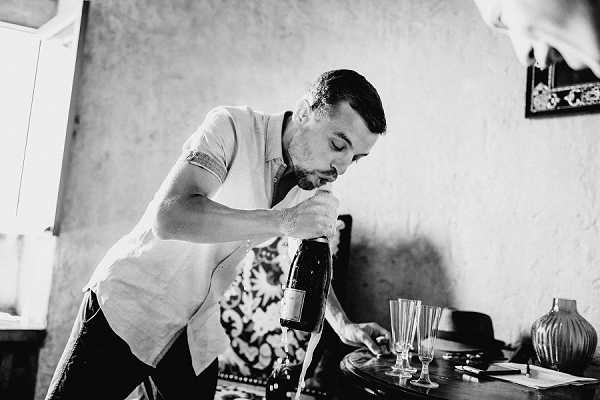 A black-and-white getting-ready moment captured indoors, showing a man leaning over a small table as he opens a large bottle of champagne with his mouth. Two empty champagne flutes and a rounded ceramic vase are visible on the table in the foreground, along with what appears to be a hat and some papers. The room has a rustic interior with textured walls and a patterned fabric visible in the background. The image is a medium close-up shot with strong contrast between the bright whites and deep shadows.