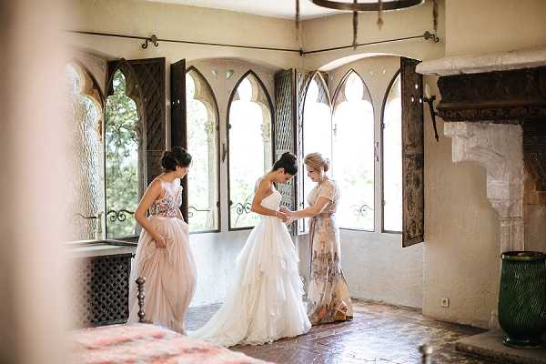 A bride is being helped into her layered ivory tulle ball gown by two women inside a historic chateau room. The bride stands in the center while one attendant on each side assists with fastening or adjusting the dress. One attendant wears a blush pink floral-embroidered gown and the other wears a short-sleeved floral printed midi dress. The room features distinctive Gothic-arched windows with dark wooden shutters, terracotta tile flooring, a large stone fireplace on the right, and a green glazed ceramic vessel in the corner, suggesting a medieval or Renaissance-era French chateau interior. The image is a medium wide shot with natural window light illuminating the scene. Potential venue feature image.