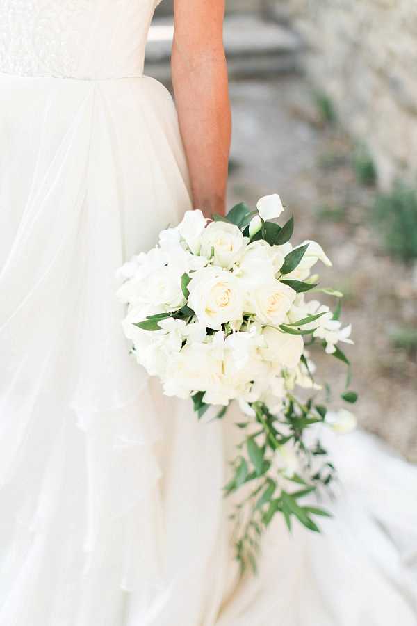 A close-up portrait of a bride holding a cascading bouquet composed of cream roses, white orchids, white freesia, and trailing green foliage including eucalyptus and fern-like stems. The bride is wearing a layered tulle ball gown with a lace bodice in ivory white. The bouquet style is a classic cascade design with a rounded top and trailing greenery dropping below the hand. The background is softly blurred, suggesting an outdoor stone setting.