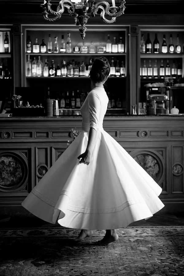 Black-and-white full-length portrait of a bride twirling alone in what appears to be an ornate interior bar area, likely within a chateau or grand hotel. She wears a structured, 1950s-inspired tea-length wedding dress with three-quarter sleeves, a fitted bodice, and a dramatically full circle skirt captured mid-spin, with visible hem detail banding near the base. Her dark hair is styled in an upswept chignon and she wears flat shoes. The background features a dark wood paneled bar with backlit shelves lined with spirit bottles, a decorative chandelier above, and an ornate patterned carpet underfoot. The image has strong contrast between the bright white of the skirt and the deep dark tones of the bar interior. Portrait shot, full figure, taken from a three-quarter rear angle.