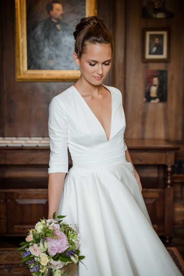 A bridal portrait shot indoors in what appears to be a wood-paneled room featuring large oil portrait paintings in ornate gold frames, suggesting a chateau or historic manor setting. The bride is photographed alone in a three-quarter portrait composition, looking downward with a relaxed expression, her dark hair styled in an upswept bun. She wears a white structured gown with a deep V-neckline, three-quarter length sleeves, and a full A-line skirt with a defined waistband, giving the dress a modern, minimalist silhouette. She holds a loosely arranged bouquet of blush pink peonies, soft ivory roses, lavender blooms, and trailing greenery.