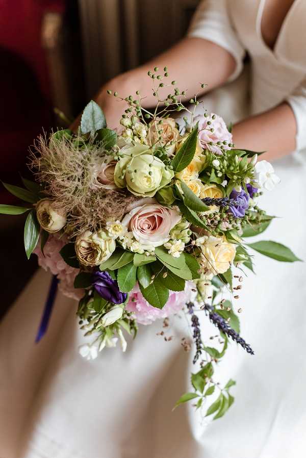 Close-up detail shot of a bride holding a loose, garden-style bridal bouquet against her ivory dress. The bouquet features blush pink roses, pale yellow spray roses, green ranunculus buds, deep purple lisianthus, light pink hydrangea, lavender stems, white small blooms, smoky thistle or clematis seed heads, unripe green berries, and abundant mixed green foliage with trailing vines. The overall color palette spans blush, cream, yellow, purple, and green, giving the arrangement a wild, organic feel consistent with a bohemian or garden-inspired styling approach. The composition is a portrait-orientation close-up focused entirely on the bouquet and the bride's torso, with a ribbon in deep purple or navy visible at the stems.