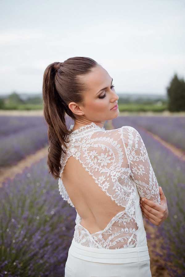 A bridal portrait shot outdoors in a lavender field in Provence, with rows of purple lavender stretching into the background. The bride is photographed from a three-quarter rear angle, showing the back detail of her white lace wedding gown, which features a deep open back with intricate floral lace panels and long sheer lace sleeves. She wears her dark brown hair in a high sleek ponytail and glances downward over her shoulder. The styling is modern and minimalist, pairing the detailed lace bodice with what appears to be a structured skirt. Close-up portrait composition with shallow depth of field blurring the lavender rows behind her.