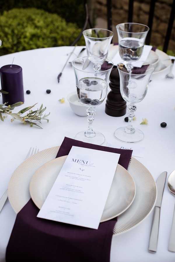 Close-up detail shot of an outdoor wedding reception table setting featuring a white tablecloth with a deep plum/aubergine color scheme. The place setting includes a gold-rimmed charger plate with a folded plum napkin and a printed white menu card on top. Multiple clear crystal wine glasses with decorative stems are arranged toward the center of the table, one containing water with floating blueberries. Scattered loose blueberries and a small olive branch sprig are used as organic table decorations. A tall deep purple pillar candle and a dark wooden pepper grinder are visible in the background. The overall styling is modern and classic with a rich jewel-tone palette of plum, gold, and white.