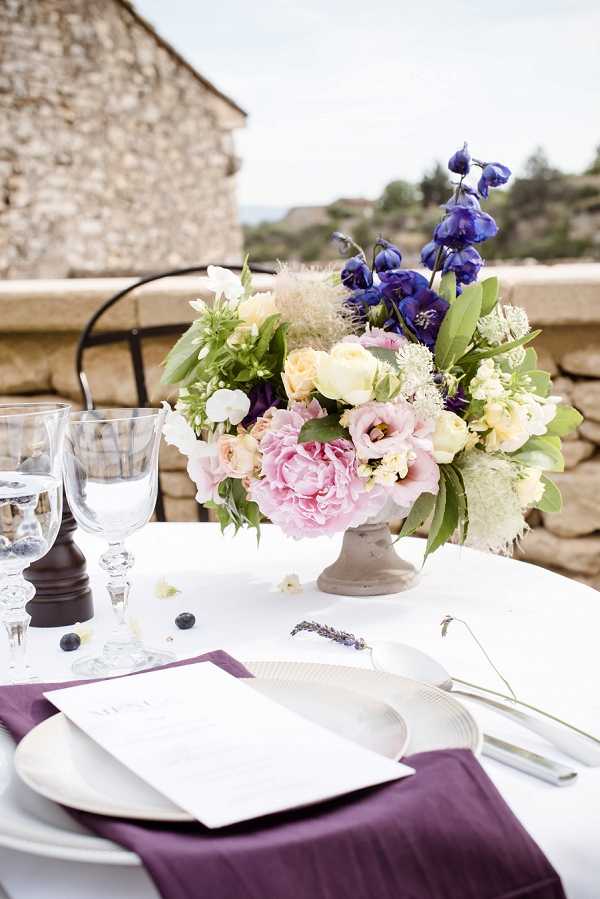 Close-up detail shot of an outdoor wedding reception table setting, likely at a stone terrace venue in the south of France. The centerpiece is a low concrete urn filled with a mixed floral arrangement featuring blush pink peonies, deep violet delphiniums, soft yellow garden roses, white lisianthus, and greenery with feathery smoke bush accents. The table is set with a white linen, deep plum/aubergine cloth napkin, a white ceramic plate, a printed menu card, silver cutlery, and crystal glassware; a few scattered blueberries and a small sprig of dried lavender serve as additional table details. The overall decor palette combines plum, blush pink, and cream tones in a classic French Provençal style.