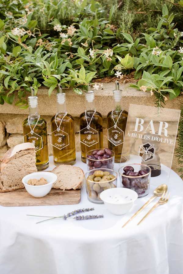 A close-up detail shot of an outdoor olive oil tasting bar ('Bar à Huiles d'Olive') set up during a cocktail hour, styled on a white linen-covered round table. Four glass bottles of infused olive oil are displayed with individual hang tags labeled with flavors including citron, lavande, and romarin, accompanied by small glass bowls of green and purple olives, a white bowl of spread, and sliced rustic bread on a wooden board. Gold cutlery and a sprig of fresh lavender are arranged in the foreground, and a modern acrylic signboard with white printed text identifies the station. The overall styling is Provençal-inspired with a clean, modern presentation.