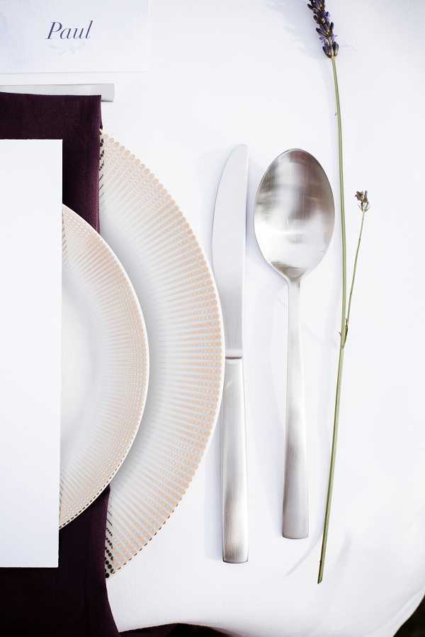 Close-up detail shot of a wedding reception place setting on a white tablecloth. The setting includes a textured cream charger plate with a radiating ribbed pattern, a white dinner plate, a deep plum/burgundy linen napkin, silver knife and spoon, a single stem of fresh lavender laid alongside the cutlery, and a white place card printed with the name 'Paul' in script font. The decor palette combines white, deep burgundy, and silver with a minimalist, modern styling approach.