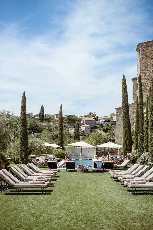 Wide shot of an outdoor pool area at a French property, likely in Provence, featuring a rectangular pool flanked by rows of beige sun loungers with rolled towels arranged symmetrically on a manicured lawn. Several large white parasols provide shade near the pool, where a small group of guests can be seen relaxing. The grounds are lined with tall cypress trees, and a historic stone building is visible on the right edge of the frame. A hillside village with terracotta-roofed buildings appears in the background. No wedding-specific activity or decor is visible in this image. Potential venue feature image.