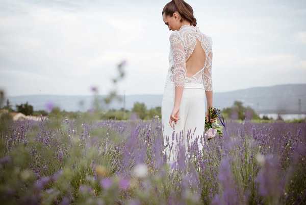 A bridal portrait taken outdoors in a lavender field, with the bride standing among rows of purple lavender in full bloom. The bride wears a two-piece white gown consisting of a fitted skirt and a three-quarter-sleeve lace top with a high neck and an open keyhole back, with intricate floral lace detailing. She holds a small bouquet featuring blush pink blooms and greenery at her side, and her dark hair is styled in a low updo. The composition is a medium shot from behind and to the side, with the lavender field filling the foreground and softly blurred hills and trees visible in the background.