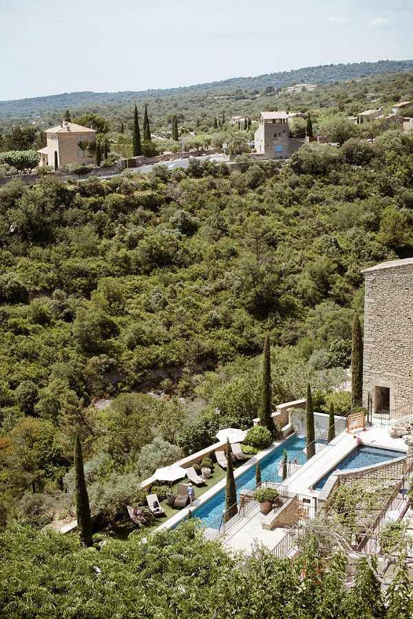 Aerial wide shot of a Provençal stone property featuring a narrow rectangular swimming pool with a surrounding terrace, white parasols, and sun loungers. The venue's natural stone architecture is visible on the right edge of the frame, with a lower terrace area below the pool level. The property is set into a hillside with dense Mediterranean vegetation and additional stone buildings visible in the background. No wedding party or ceremony activity is visible in this shot. Potential venue feature image.