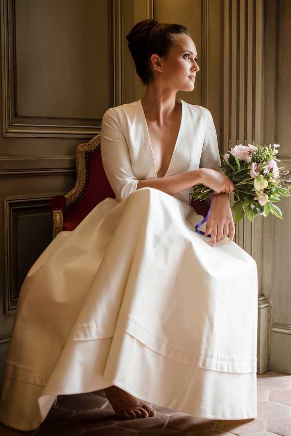 A bridal portrait of a single bride seated in a red and gold carved armchair inside a room with panelled grey-green walls, likely within a French chateau or manor house. She wears a minimalist ivory satin gown with a deep V-neckline and three-quarter-length sleeves, with a full tiered skirt, and is barefoot. She holds a loose, garden-style bouquet of blush pink peonies, soft lavender blooms, and green foliage tied with a cobalt blue ribbon, and gazes off to the side. The composition is a medium portrait shot with warm, diffused natural light coming from the side.