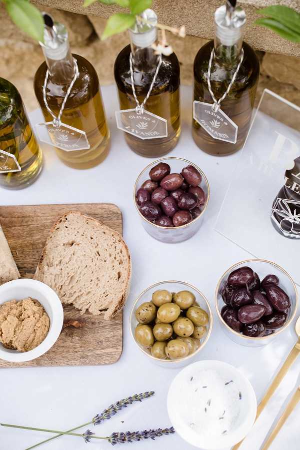 Close-up detail shot of a cocktail hour food station set on a white linen-covered table. Three glass bottles of infused olive oil are displayed at the back, each tagged with printed labels reading 'Olives & Lavande' and similar French text, tied with twine. In front of the bottles are three gold-rimmed glass bowls filled with black Kalamata olives and green olives respectively. A wooden cutting board holds sliced rustic bread and a small white bowl of tapenade or spread. A small white dish in the foreground contains a dip garnished with dried lavender seeds, and two sprigs of fresh lavender are placed decoratively on the table surface. The styling has a Provençal theme with a simple, clean palette of white, gold, and natural wood tones.