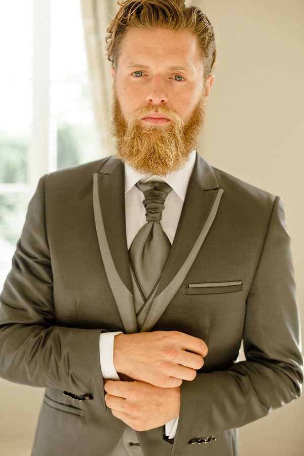 Close-up portrait of a groom getting ready, shot indoors against a neutral beige background with soft natural window light. The groom has auburn hair and a full red-brown beard, and is adjusting his cufflinks. He is wearing a charcoal grey suit with satin shawl lapels and a matching grey waistcoat, paired with a white dress shirt and a charcoal grey silk cravat tied in an ascot knot. The styling is classic and formal, with a monochromatic grey palette throughout the outfit.