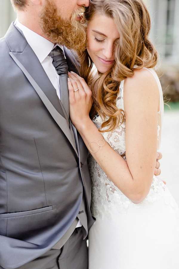 A close-up couple portrait showing the bride leaning into the groom with her eyes closed and a soft smile, her hand resting on his chest near his tie. The groom, who has a full red beard, wears a light grey suit with a charcoal grey tie and white dress shirt. The bride wears a backless lace gown with floral appliqué detailing and a delicate circular-link bracelet, with long wavy auburn hair worn down. The composition is a tight mid-shot cropping the groom's face and focusing on the couple's connection, with a softly blurred architectural background.