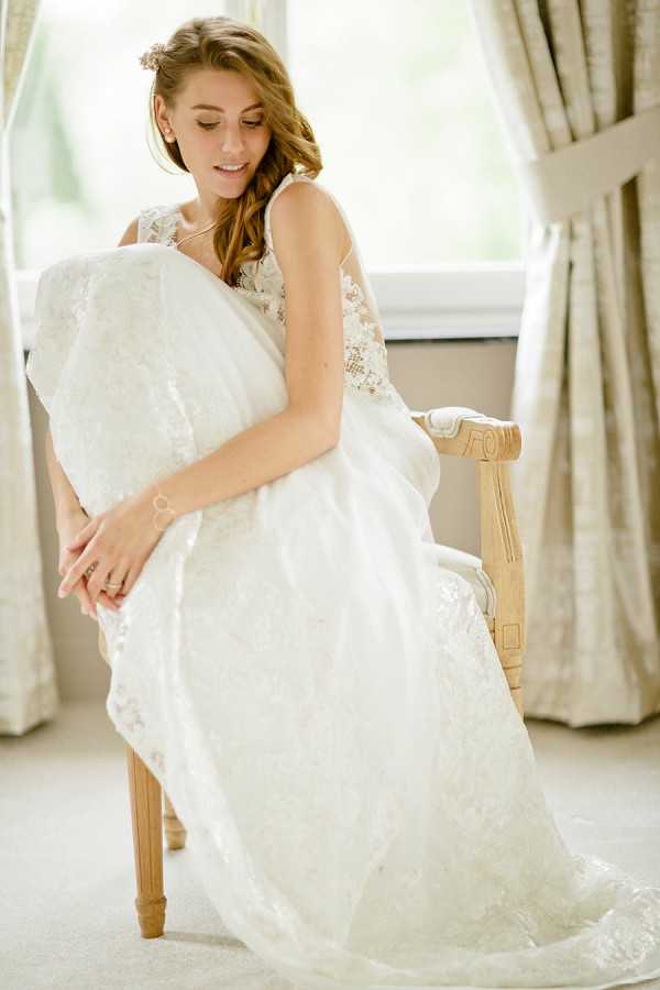 A bridal portrait taken indoors during the getting-ready phase, showing a bride seated on a light natural wood armchair near a window with neutral floral curtains. She is wearing an ivory lace wedding gown with a lace-appliqué bodice and illusion back detailing, with the full skirt and train pooling around the chair. Her auburn hair is worn in loose waves with a small floral hair accessory, and she is looking downward with a relaxed expression. The image is a medium-full portrait shot with soft, diffused natural window light creating a bright, airy feel.