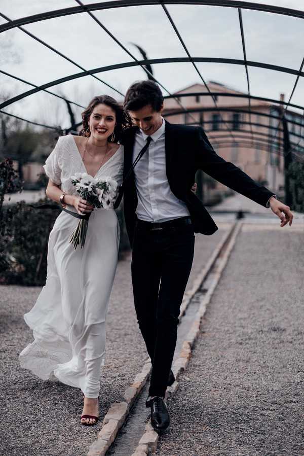 A couple portrait taken outdoors beneath a curved metal pergola structure on a gravel path, with a large historic building visible in the background. The bride wears a flowing white wrap-style dress with short sleeves and a deep V-neckline, paired with dark burgundy heeled sandals and bold red lipstick, and carries a small bouquet of white flowers with minimal greenery. The groom wears a black suit with a white shirt and a loosened black tie, dressed in black oxford shoes. The two are walking and laughing together, with the groom playfully extending one arm outward, captured in a candid full-length portrait shot with a modern, editorial styling aesthetic.