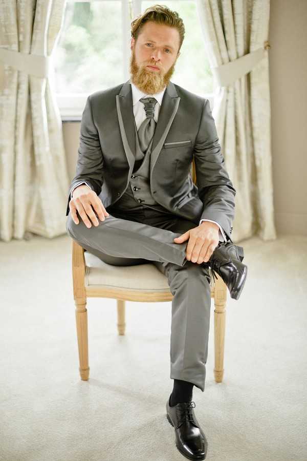 A portrait of the groom seated on a wooden chair with a cream upholstered seat, photographed indoors in front of a window with cream curtains. He is wearing a charcoal grey three-piece suit with satin lapel trim, a white dress shirt, a grey textured tie, black dress socks, and black oxford shoes, and holds one shoe in his hand in a relaxed pose. The groom has a full auburn beard and styled hair, and looks directly at the camera. The shot is a mid-length portrait with soft natural window light.